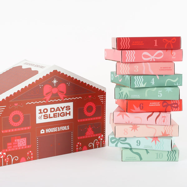 10 Days of Sleigh gingerbread house advent calendar beside a stack of ten numbered mystery foil boxes